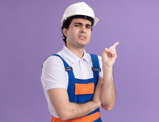 混乱年轻的建筑工人穿着建筑制服 戴着安全帽 看上去很困惑 用食指指着站在紫色墙上的一边手指头盔制服