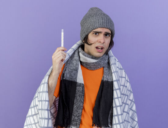格子布忧心忡忡的年轻人戴着冬天的帽子 围巾裹着格子呢 手里拿着隔离在紫色背景上的温度计抱着关心疾病