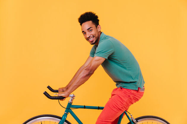 着装运动型的非洲黑发男人在自行车上摆姿势快乐的微笑的家伙在到处游荡休闲时髦自行车