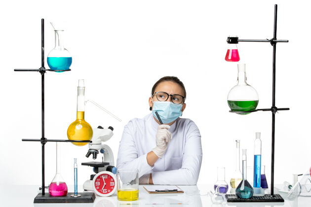 前面前视图穿着医疗服的女化学家 戴着面罩 拿着溶液坐着 在白色背景上写笔记 溅起实验室病毒化学冠状病毒笔记喷溅医学