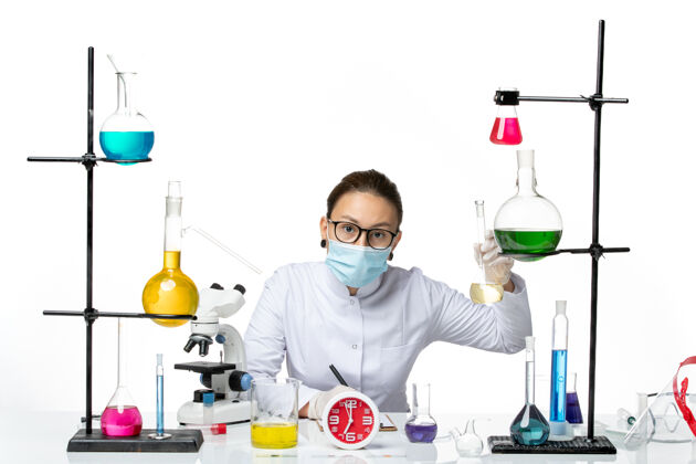 实验室前视图：穿着白色医疗服的女化学家 带着面罩 桌子前面有白色背景上的溶液化学家实验室的病毒病毒病毒喷溅实验室专业科学