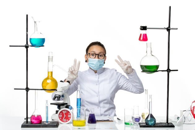 病毒正面图：穿着医疗服的女化学家 戴着口罩坐在桌子前面 白色桌子上有解决方案病毒化学实验室covidsplash实验室科学解决方案
