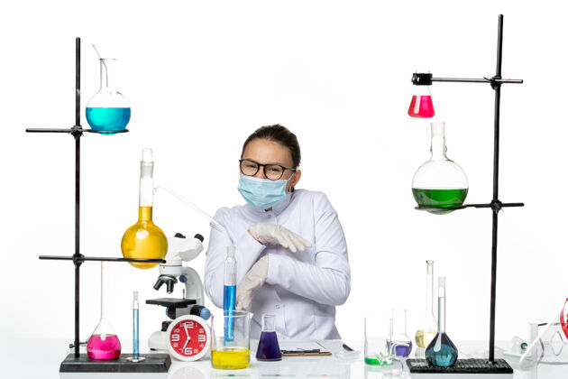 病毒前视图穿着医疗服的女化学家带着面罩坐在桌子前面 浅白的背景上是病毒化学实验室的病毒飞溅医学化学坐着