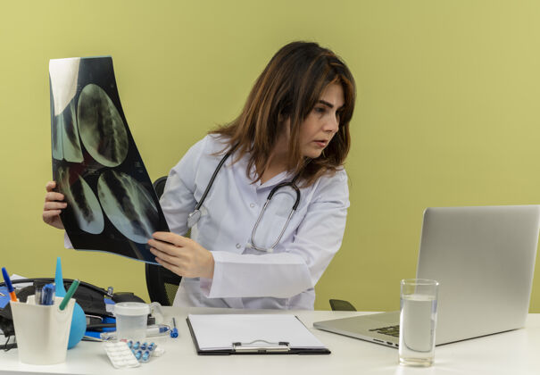 X光集中中年女医生穿着医用长袍和听诊器坐在办公桌前拿着医用工具拿着x光拍看着笔记本电脑坐着医疗抱着