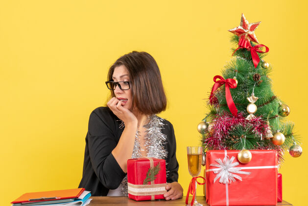 微笑好奇的商务女士穿着西装 戴着眼镜 手里拿着礼物 低头坐在一张桌子旁 桌子上放着一棵圣诞树快乐好奇坐着