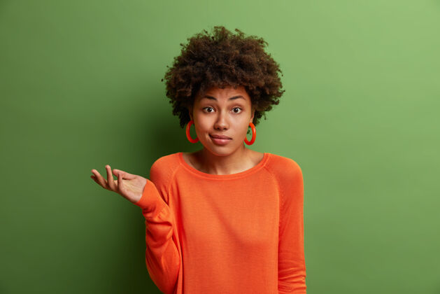 反应犹豫不决的女人迟疑地举起手掌 面对难题或两个选择 穿着橙色毛衣和耳环 孤零零地站在绿色的墙上人 感知和态度种族成人怀疑