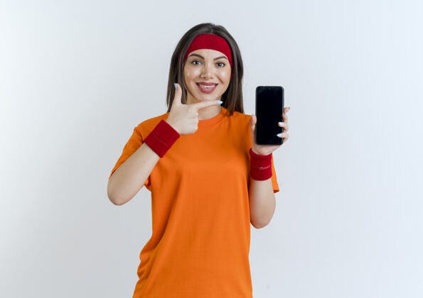 头带微笑的年轻运动女性戴着头带和腕带显示手机指着它看起来孤立显示电话人