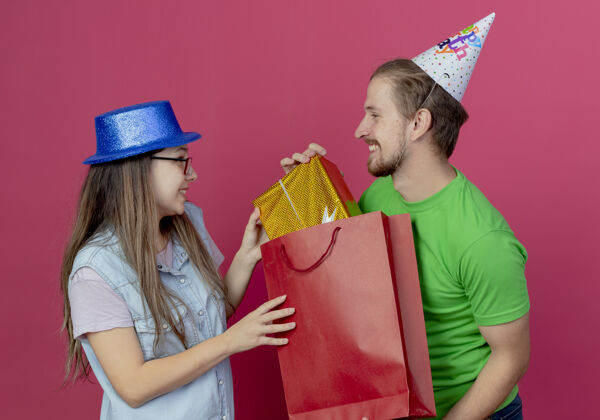盒子一对戴着派对礼帽的年轻夫妇看着对方 从粉色墙上的红色购物袋里掏出礼盒生日男人红色