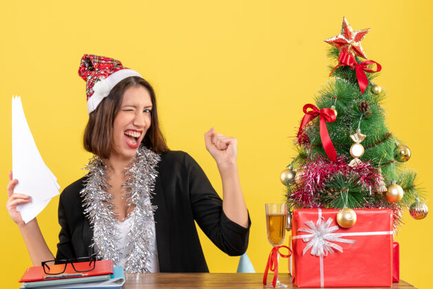 花束情绪化的商务女士穿着西装 戴着圣诞老人帽和新年装饰品 手里拿着文件 坐在一张桌子旁 桌子上放着圣诞树桌子礼物漂亮