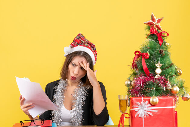 困惑一个穿着西装 戴着圣诞老人帽 戴着新年装饰品 手里拿着文件 坐在一张桌子旁 桌子上放着圣诞树的困惑的商务女士举行桌子性感