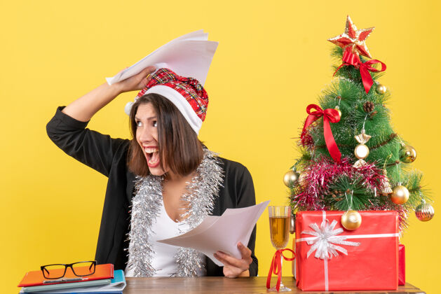 漂亮情绪疲惫的商务女士穿着西装 戴着圣诞老人帽和新年装饰品 手里拿着文件 坐在一张桌子旁 桌子上放着圣诞树抱着圣诞老人树