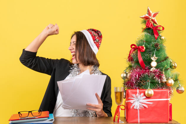 帽子情绪高昂的商务女士穿着西装 戴着圣诞老人的帽子和新年装饰品 手里拿着文件 坐在一张桌子旁 桌子上放着圣诞树文件自豪圣诞老人