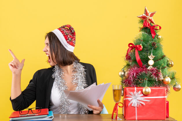 文件情绪紧张的商务女士穿着西装 戴着圣诞老人帽和新年装饰品 手里拿着文件 坐在一张桌子旁 桌子上放着圣诞树年份树帽子