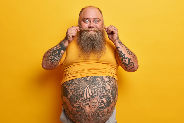 腹部橫拍搞笑的胖家伙 大肚子 手臂和腹部紋身 扭胡子 穿著黃色t恤 肥胖 因為喝了很多啤酒 吃垃圾食品胖懶男年輕體重陽剛
