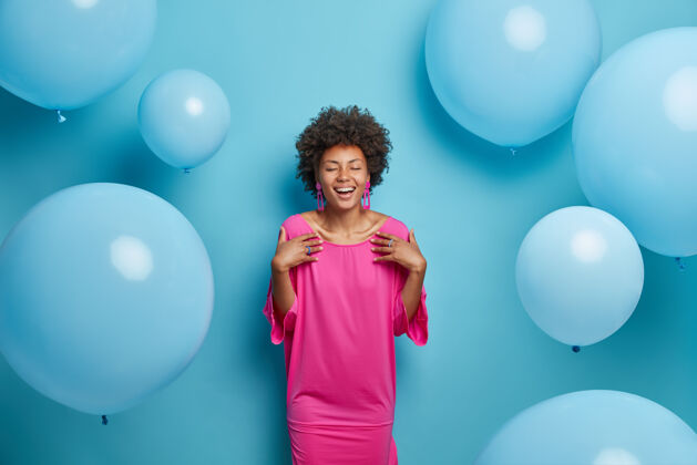 连衣裙室内拍摄了一位身着粉色时装的快乐卷发女士 闭上眼睛 准备特殊场合 高兴地用生日来迎接祝贺 孤立在蓝色背景上 充气气球氦衣服情绪