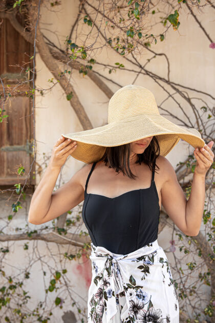 休息一个年轻的女人躲在一个大草帽下躲避太阳 草帽靠近一堵墙 墙上缠绕着一棵正在开花的干树枝时尚乡村帽子
