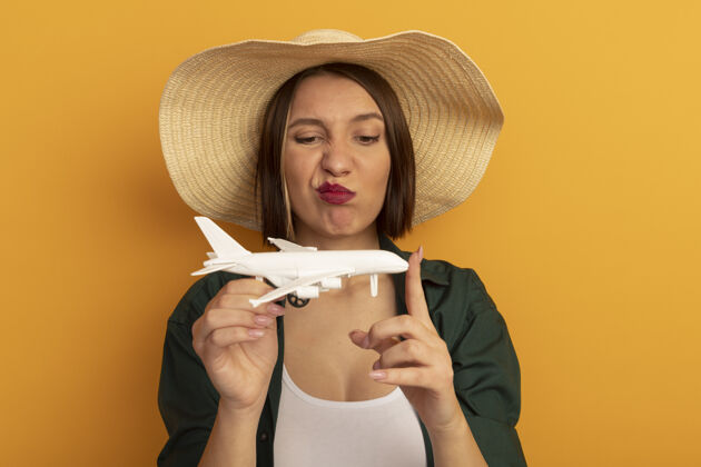 脸一个戴着沙滩帽的漂亮女人看着隔离在橘色墙上的飞机模型漂亮站着帽子