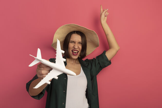 举行戴着沙滩帽的快乐美女手持飞机模型 孤零零地站在粉红色的墙上漂亮人模特