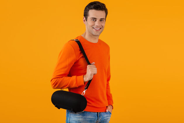 便携时尚微笑的年轻人穿着橙色毛衣手持无线扬声器快乐地听音乐无线举行设备