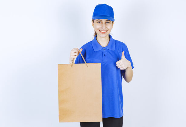 服务穿着蓝色制服的女孩手里拿着一个纸板购物袋 展示着满意的标志积极人模特