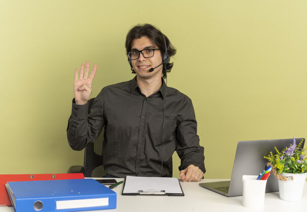 笔记本电脑戴着耳机 戴着眼镜 面带微笑的年轻上班族坐在办公桌旁 拿着办公工具 用笔记本电脑做手势 绿色背景上有四个手势 并留有复印空间耳机四手