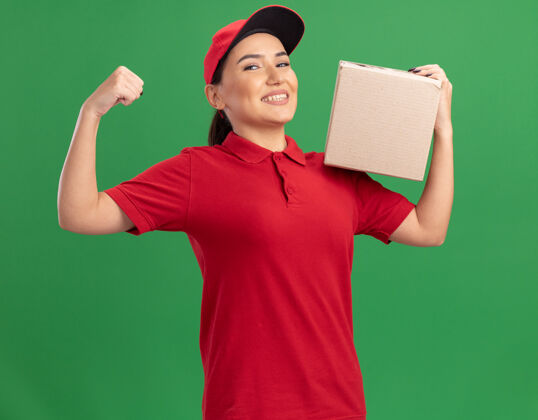 喜欢身着红色制服 头戴鸭舌帽 手持纸箱 面带微笑 高举拳头的快乐年轻女送货员站在绿色的墙壁上交货纸板盒子