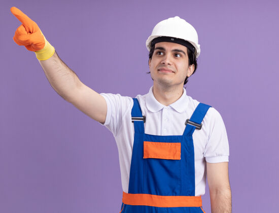橡胶年轻的建筑工人穿着建筑制服 戴着安全帽 戴着橡胶手套 用食指指着紫色墙上站着的微笑的东西制服目录微笑