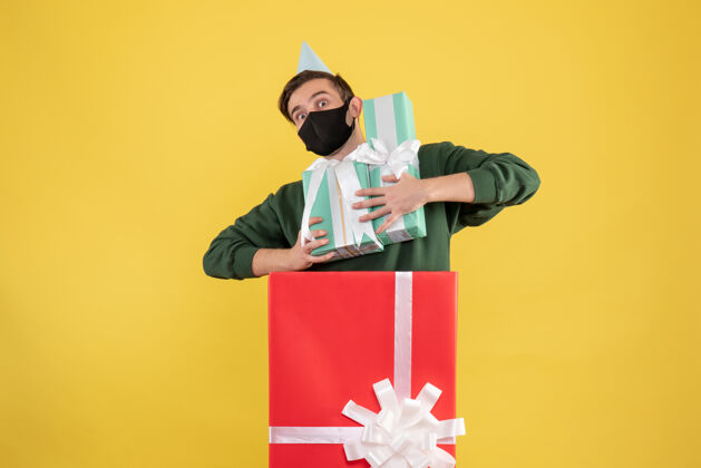 背景正面图戴着派对帽的年轻人拿着圣诞礼物站在黄色背景的大礼盒后面成人前面立场