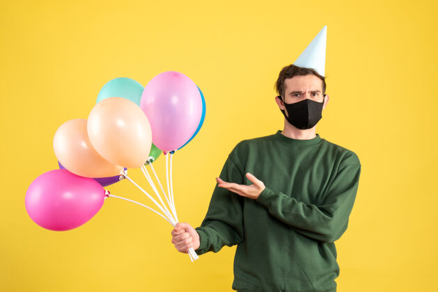 快乐正面图：戴着派对帽 站在黄色背景上的彩色气球的年轻人氧气前面帽子