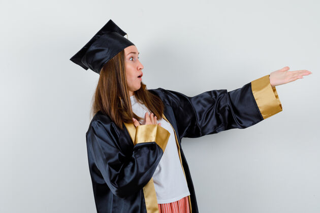 成人女毕业生手放在胸前 穿着学院服伸着手 看上去很惊讶正面图保持肖像伸展