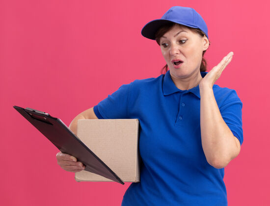 站着身穿蓝色制服 戴着帽子的中年女送货员拿着纸板箱 站在粉红色的墙上 困惑地看着剪贴板硬纸板年龄拿着