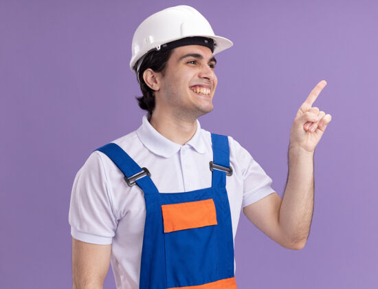 建筑年轻的建筑工人穿着建筑制服 戴着安全帽 一边微笑着用食指指着紫色墙壁上的东西头盔制服手指