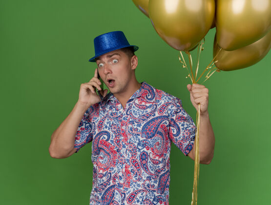 气球惊讶的年轻人戴着蓝色帽子拿着气球在电话里讲话绿色背景隔离说话戴着惊喜