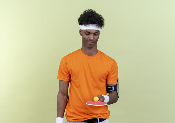 球拍高兴的年轻黑人美国运动男子戴着头带和腕带举行 看着乒乓球拍与球隔离在绿色的背景腕带持有球