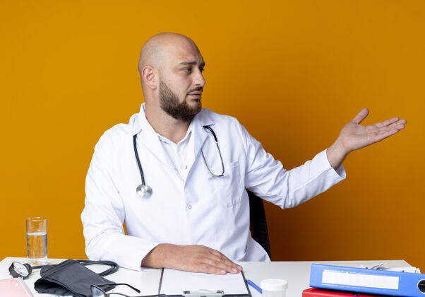 男性看着身边年轻秃顶的男医生穿着医用长袍和听诊器坐在办公桌前 用医用工具指着隔离在橙色背景上的一侧工具办公桌听诊器