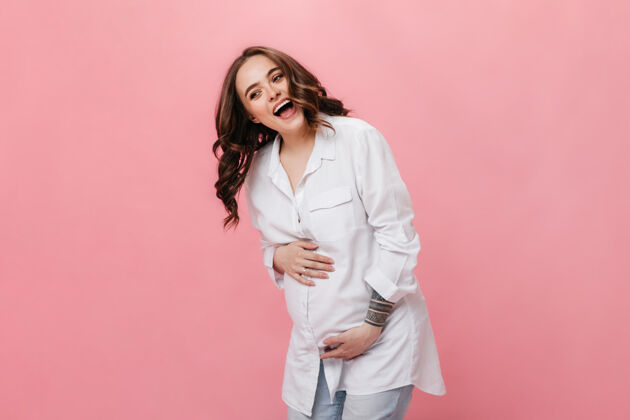 怀孕穿着白色长衫的情绪化快乐的女人的肖像穿着牛仔裤的快乐孕妇在粉色背景上笑着摆姿势牛仔裤室内怀孕
