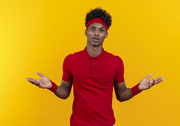 运动困惑的年轻黑人美国运动男子戴着头带和腕带展开双手孤立在黄色的背景蔓延非裔美国人年轻
