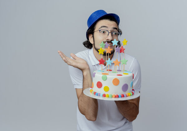 蛋糕看着身边迷茫的帅哥戴着眼镜和蓝帽子拿着蛋糕 摊开手隔离在白色的背景上摊开戴着帽子