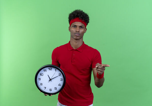 运动自信的美国黑人运动型年轻人戴着头带和腕带 手持挂钟 在绿色背景上向你展示孤立的手势腕带墙穿