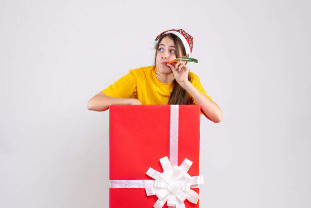 人前视图好奇的女孩 戴着圣诞帽 站在圣诞礼物后面 用隔音器快乐使用爱国
