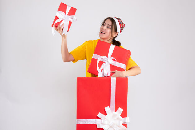 购物者正面图可爱的圣诞女孩戴着圣诞帽拿着礼物站在大圣诞礼物后面漂亮帽子立场
