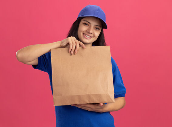 帽子身着蓝色制服 头戴礼帽 面带微笑 站在粉色墙壁上的快乐年轻送货女孩站立包装表演