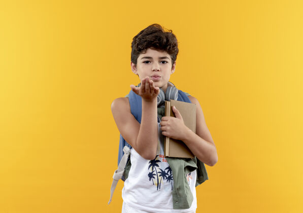 抱看着镜头 一个背着书包 戴着耳机 拿着书 在黄色背景下展示亲吻动作的小男孩吻包学校