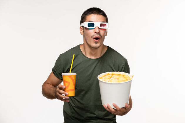电影正面图身穿绿色t恤的年轻男性手持土豆cips和苏打水 戴着d太阳镜 在白色墙上拍摄男性孤独电影土豆果汁咖啡