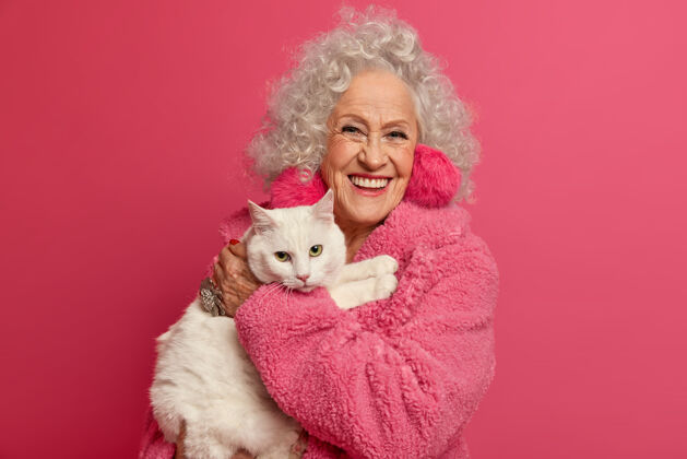 养老金满脸皱纹的祖母手牵白猫 大流行期间呆在家里 戴着蓬松的耳环 穿着柔软的长袍 准备喂宠物 隔离在粉红色的墙上女人领养老金主人朋友猫