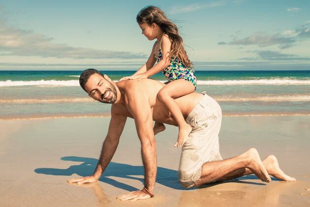侧视快乐的爸爸双手跪在沙滩上 背着小女孩自由享受蓝色