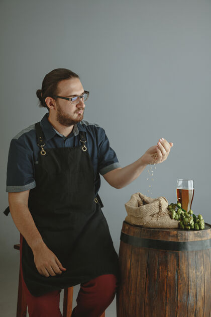 年轻人自信的年轻男性酿酒师 在灰色的木桶上用玻璃瓶自制啤酒人木材工厂