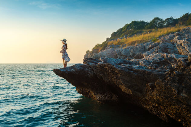 成功站在岩石顶上 望着四长岛的海边和日落的年轻女子山顶顶部帽子