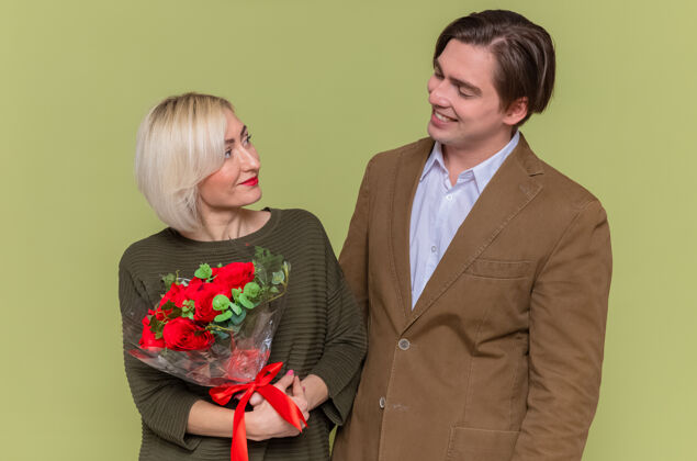 三月年轻漂亮的夫妇幸福的男人看着他可爱的女朋友和一束红玫瑰站在绿色的墙上庆祝国际妇女节情侣站庆祝