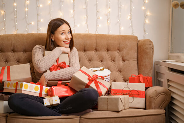 女人幸福的年轻女子抱着许多礼物盒盘腿坐在一个驼色沙发与灯女性圣诞节年轻
